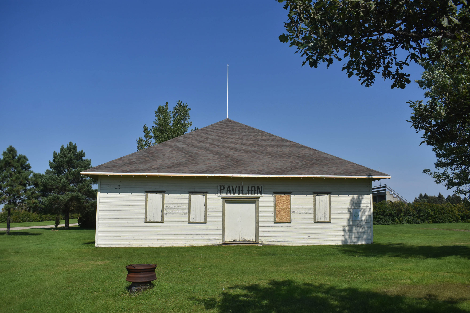 White wooden building with four wood covered windows and wooden doors. There's a big white pole sticking up from the peak of theroof. PAVILION is written above the doors.