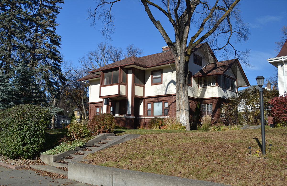Exterior shot of a two-story house with the lower level being brown and the upper level being tan with brown trim. The roof is also brown. A cement staircase leads up to the house from the sidewalk.