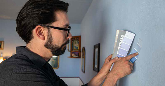 A man with dark hair and glasses holds up blue/gray paint swatches next to a blue/gray wall to match them